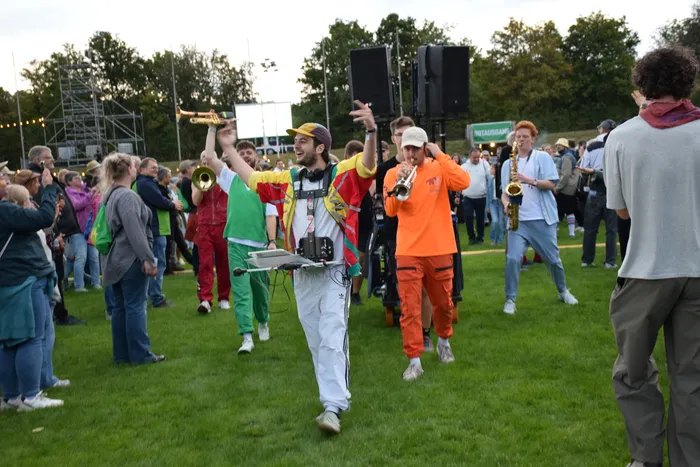 Musiker in farbenfroher Kleidung spielen Blasinstrumente inmitten einer Menschenmenge auf einer Wiese.