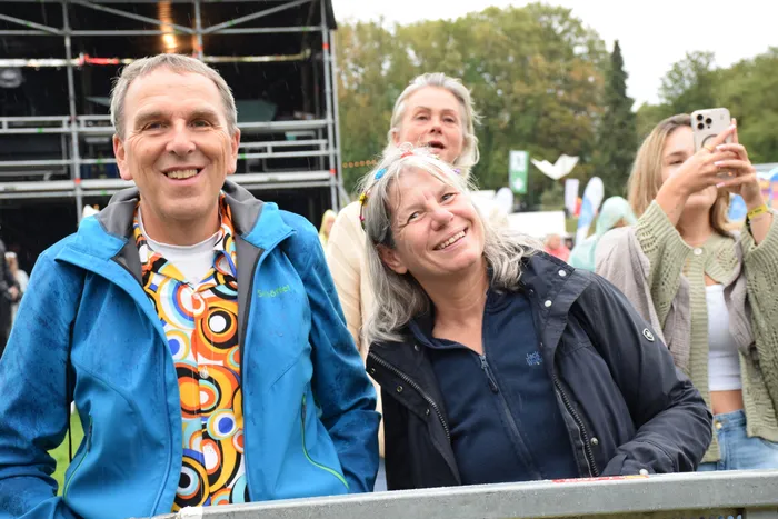 Ein lächelnder Mann in blauer Jacke und eine Frau in dunkler Jacke stehen hinter einer Absperrung bei einer Open-Air-Veranstaltung.