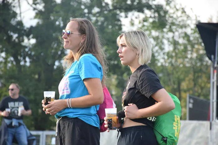 Zwei junge Frauen mit Getränken in den Händen stehen bei einem Sommerfestival im Freien.