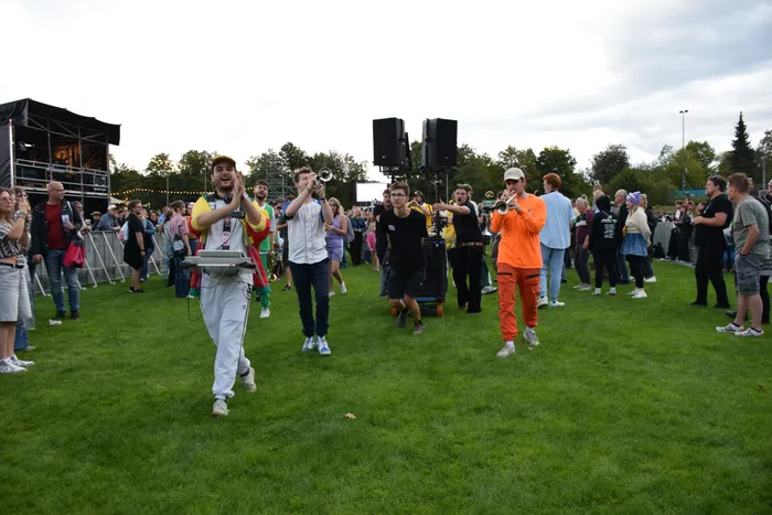 Musiker einer Brassband spielen Blasinstrumente und Perkussion inmitten einer Menschenmenge auf einer Wiese.