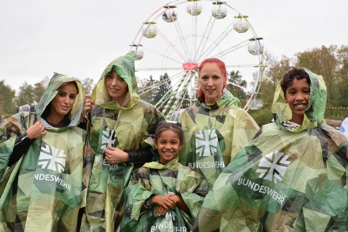 Vier lächelnde Personen in grün-gemusterten Bundeswehr-Regenponchos vor einem Riesenrad.