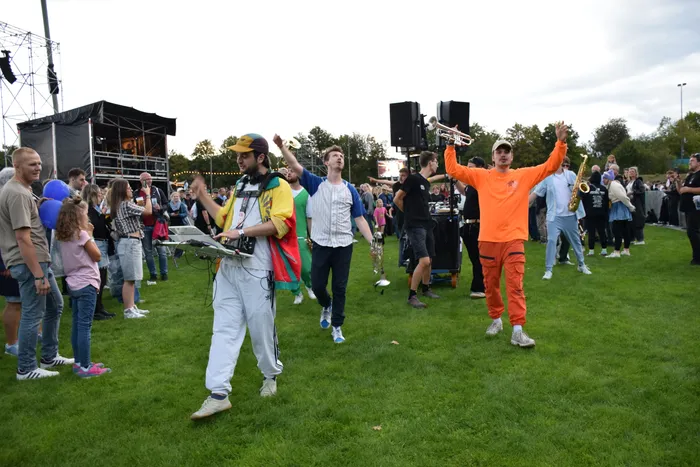 Musiker einer Brassband in bunter Kleidung spielen Instrumente wie Trompete und Keyboard inmitten einer Menschenmenge auf einer Wiese.