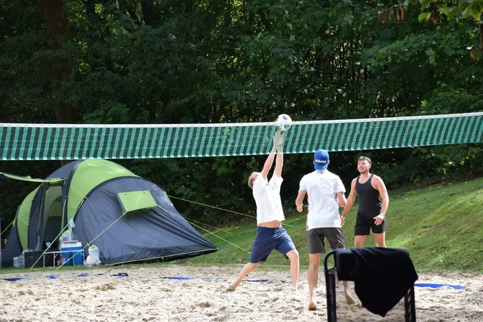 Ein Mann springt auf einem Sandplatz hoch, um einen Volleyball über das Netz zu spielen. Im Hintergrund stehen Bäume und ein grün-graues Zelt.
