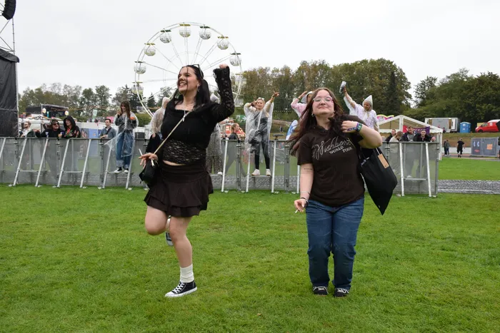 Zwei junge Frauen tanzen ausgelassen auf einer Wiese vor einem Riesenrad bei einem Festival.