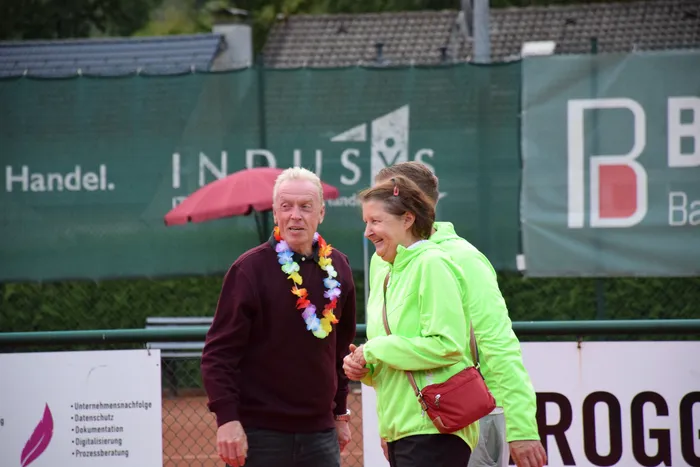 Ein Mann mit einer bunten Blumenkette und eine Frau in einer neongrünen Jacke lächeln auf einer Tennisanlage.