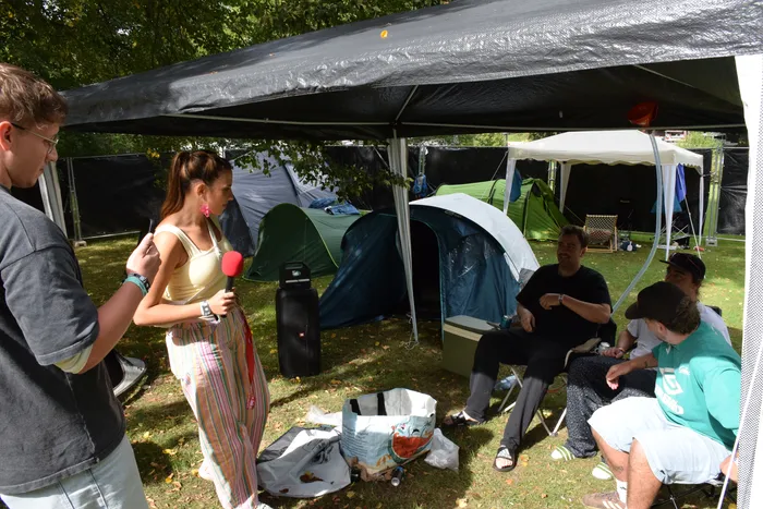 Eine Frau wird unter einem schwarzen Zeltdach mit einem roten Mikrofon interviewt, im Hintergrund stehen mehrere Campingzelte.