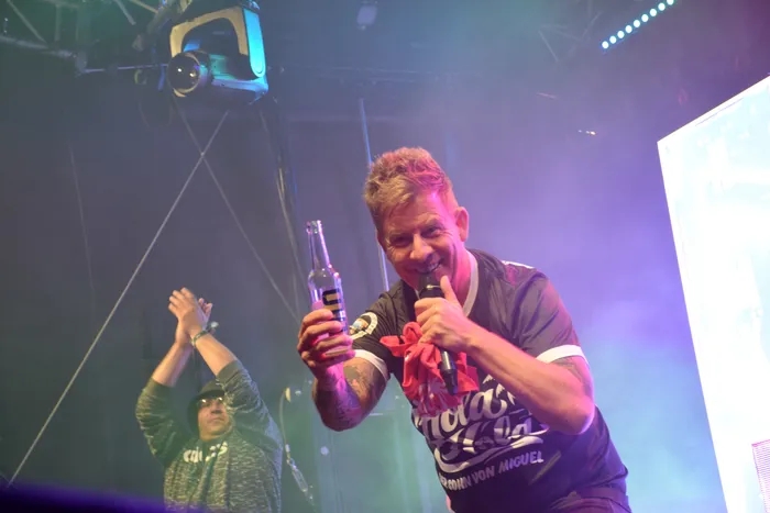 Almklausi lacht auf der Bühne beim Bautz 2026, während er eine Bierflasche und ein Mikrofon hält.