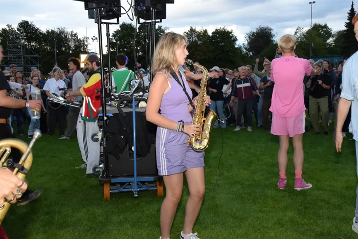 Eine junge Frau in lila Kleidung spielt Saxophon inmitten einer Menschenmenge auf einer Rasenfläche.