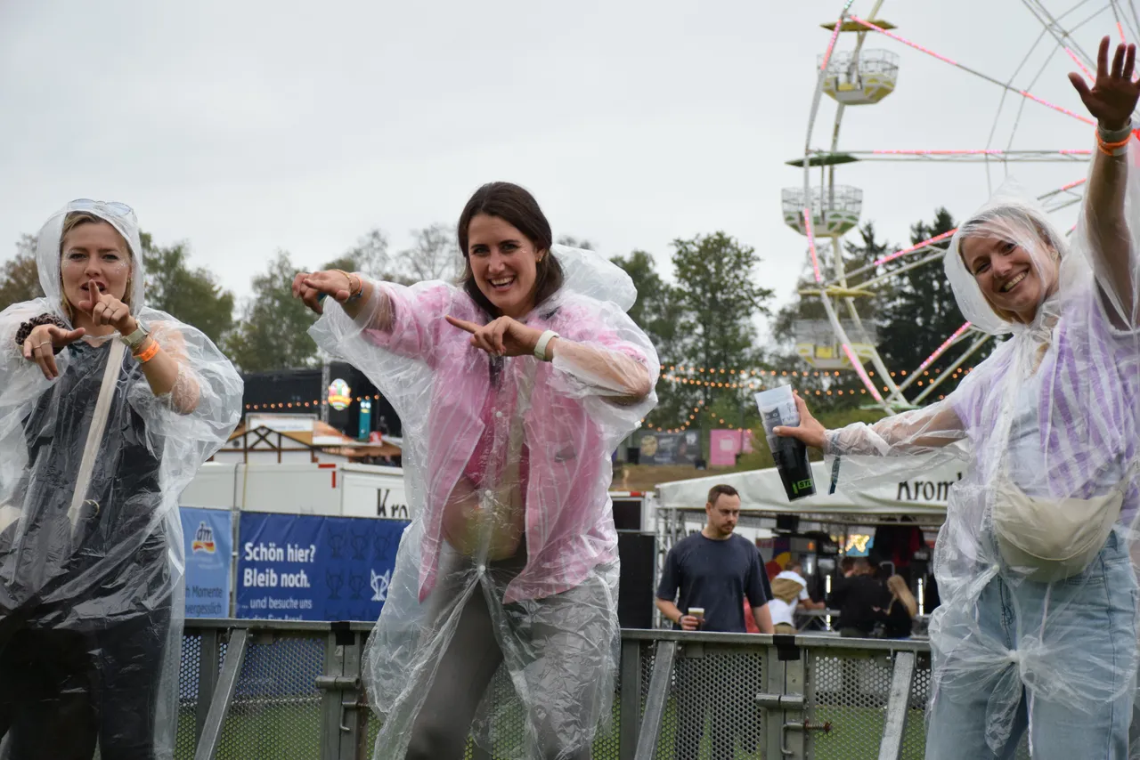 Drei Frauen in transparenten Regenponchos tanzen ausgelassen vor einem Riesenrad bei einem Open-Air-Event.