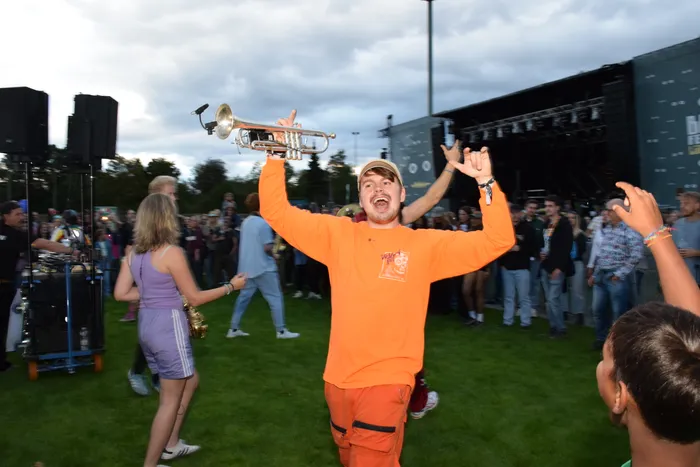 Ein jubelnder Mann in orangefarbener Kleidung hält eine Trompete hoch in die Luft inmitten einer Menschenmenge vor einer Bühne.