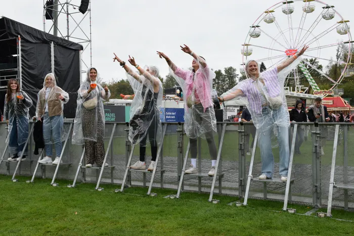 Eine Gruppe von Frauen in transparenten Regenmänteln tanzt ausgelassen auf Metallstufen vor einem Riesenrad.