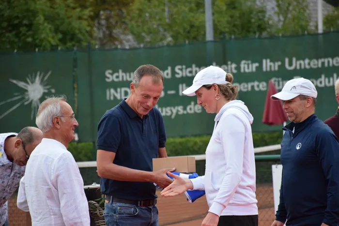 Ein Mann in einem dunkelblauen Poloshirt überreicht einer Frau in weißer Kleidung eine braune Schachtel bei einer Jubiläumsfeier auf einer Tennisanlage.
