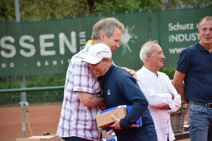 Zwei Männer umarmen sich bei der Jubiläumsfeier der MTC/TC Halver Open auf einer Tennisanlage; einer trägt eine Blumenkette.