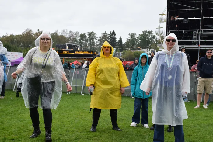 Besucher in transparenten, gelben und blauen Regenponchos stehen auf einer Wiese vor einer Bühne.