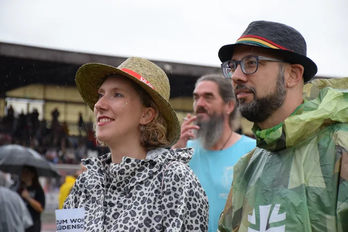 Eine lächelnde Frau mit Strohhut und Leopardenmuster-Regenjacke steht neben einem Mann mit schwarzem Hut und grüner Camouflage-Regenjacke.