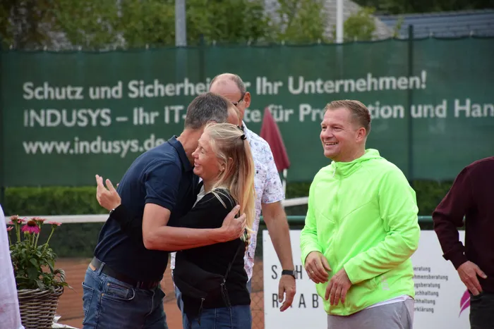 Besucher umarmen sich und lachen bei der Feier zum 20-jährigen Jubiläum der MTC/TC Halver Open auf einer Tennisanlage.