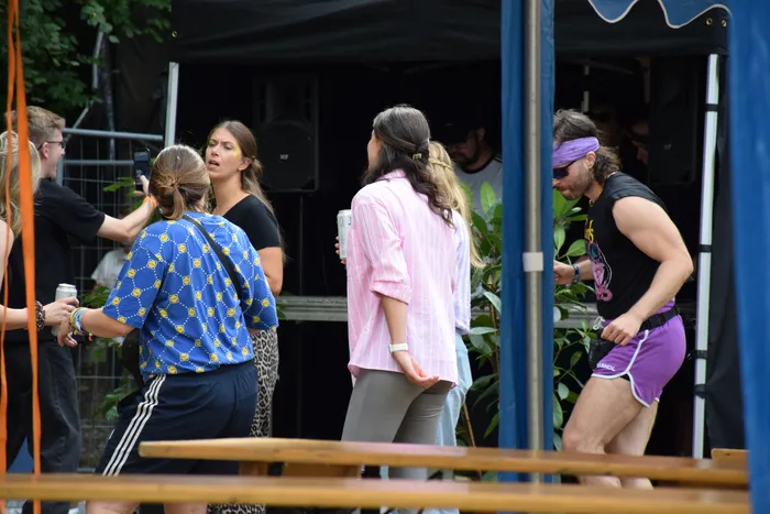 Menschen stehen bei einer Veranstaltung im Freien vor einer Bühne, darunter eine Frau in einem blau gemusterten Hemd und ein Mann in einem schwarzen Tanktop und lila Shorts.