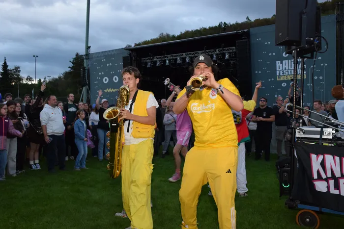 Zwei Musiker in gelber Kleidung spielen Saxophon und Trompete vor einer Menschenmenge bei einem Festival.