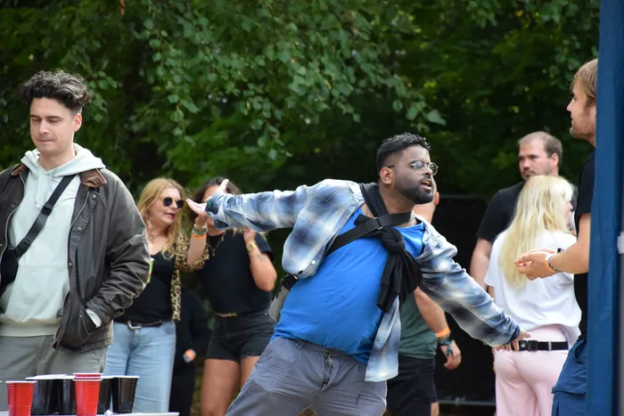 Ein Mann in einem blauen T-Shirt und karierter Hemdjacke tanzt mit ausgestreckten Armen vor einer Gruppe von Menschen im Freien.