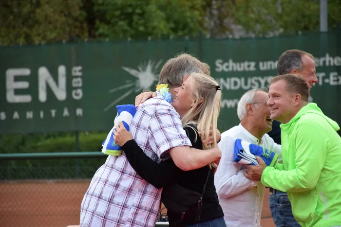 Ein Paar umarmt sich bei der Jubiläumsfeier der MTC/TC Halver Open, während im Hintergrund lachende Menschen stehen.