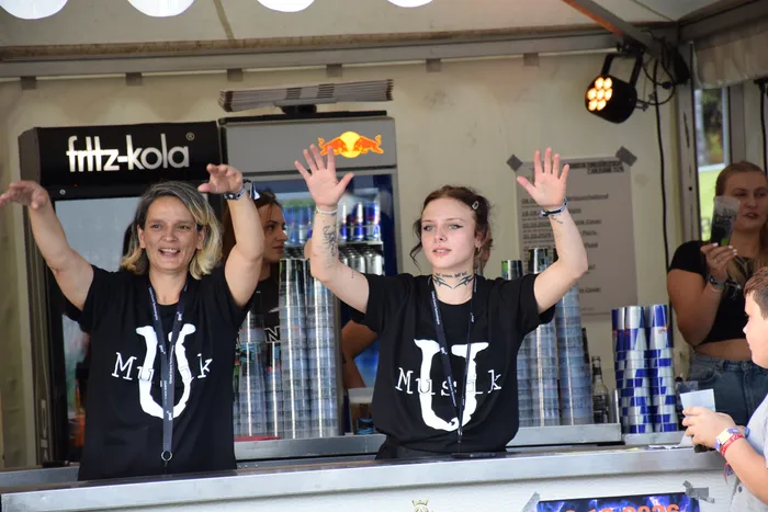 Zwei Frauen in schwarzen T-Shirts mit weißem „Musik“-Aufdruck heben jubelnd die Hände hinter einem Verkaufstresen.
