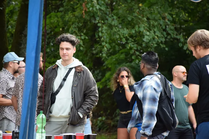 Ein Mann in einer braunen Jacke und einem weißen Kapuzenpullover steht im Vordergrund einer Gruppe von Menschen bei einem Outdoor-Event im Grünen.