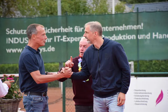 Zwei Männer schütteln sich auf einem Tennisplatz die Hände, während ein dritter Mann mit einer bunten Blumenkette im Hintergrund steht.