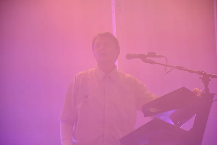 Ein Musiker steht hinter einem Keyboard und einem Mikrofon in pinkes Bühnenlicht getaucht.