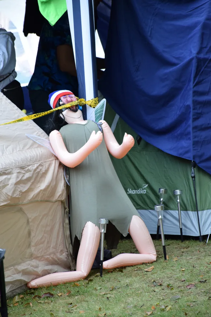 Aufblasbare Figur mit Flügeln in einem grünen Kleid und einem rot-weiß-blauen Hut zwischen zwei Zelten auf einer Wiese.