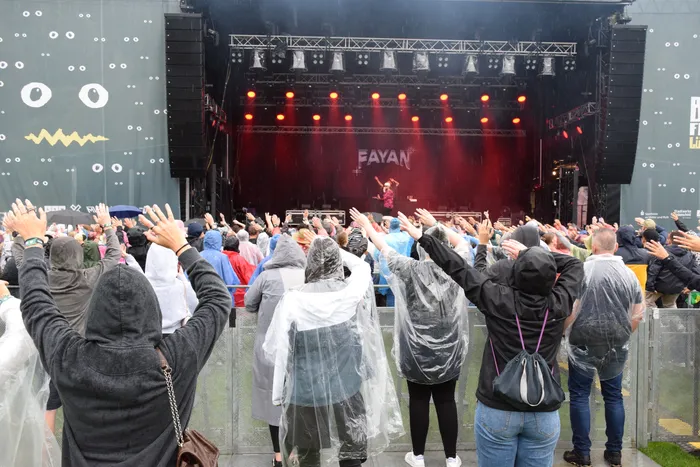 Eine Menge von Zuschauern in Regenbekleidung mit erhobenen Händen vor einer rot beleuchteten Bühne mit dem Schriftzug „FAYAN“.