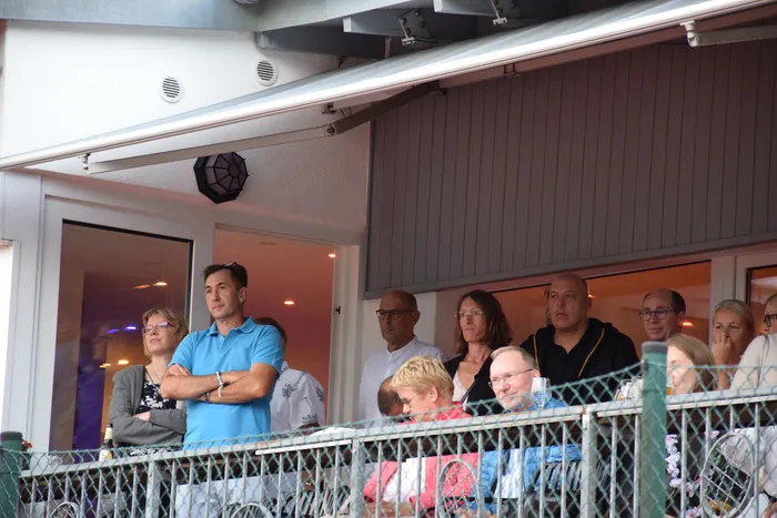 Eine Gruppe von Menschen steht bei der Jubiläumsfeier der MTC/TC Halver Open hinter einem Metallzaun auf einer Tennisanlage.