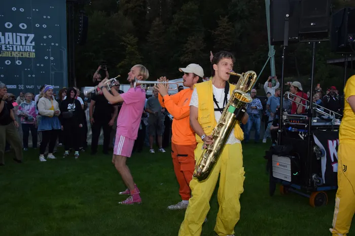 Musiker in leuchtend farbigen Outfits spielen Trompeten und Saxophon vor einer Menschenmenge bei einem Festival.