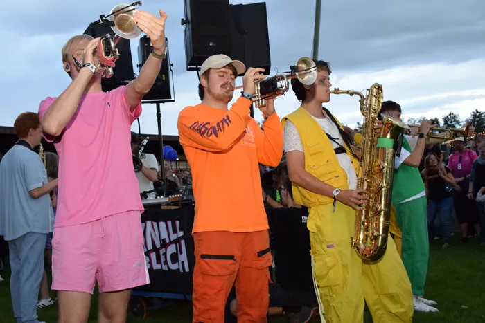 Musiker in leuchtend pinken, orangefarbenen und gelben Outfits spielen Trompete und Saxophon im Freien.