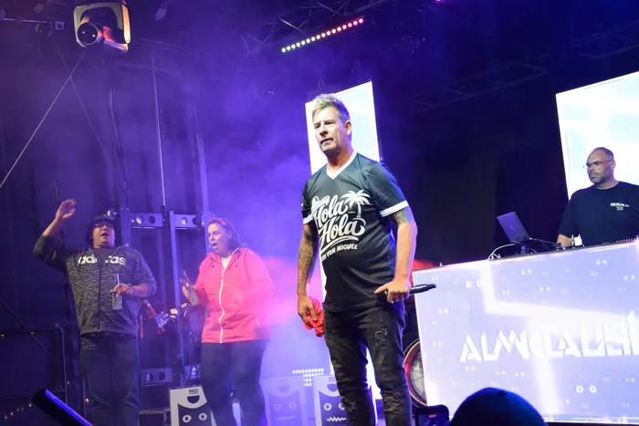 Almklausi performt auf einer Bühne unter lila Scheinwerferlicht und trägt ein schwarzes T-Shirt mit Palmenmotiv und der Aufschrift „Hola Hola“.