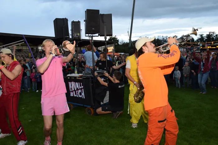 Mitglieder der Band Knallblech spielen Blasinstrumente inmitten einer Menschenmenge bei einer Open-Air-Veranstaltung.