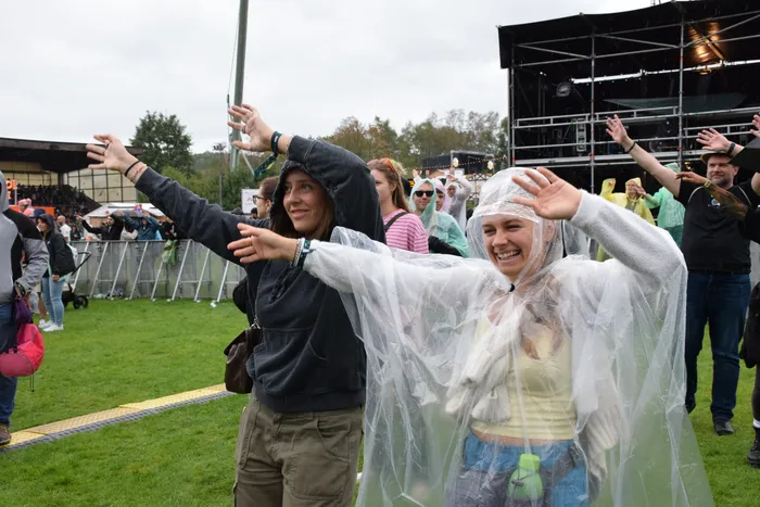 Eine lachende Frau in einem durchsichtigen Regenmantel und eine weitere Frau mit erhobenen Armen feiern bei einem Festival im Regen.