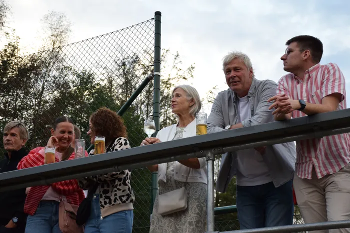 Mehrere Personen stehen mit Getränken hinter einem Geländer und blicken auf die Feierlichkeiten der MTC/TC Halver Open auf einer Tennisanlage.