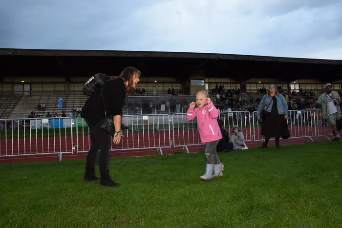 Ein Kind in einer rosa Jacke springt auf einer Wiese, während eine Frau daneben lacht. Im Hintergrund sind eine Laufbahn und Stadiontribünen zu sehen.