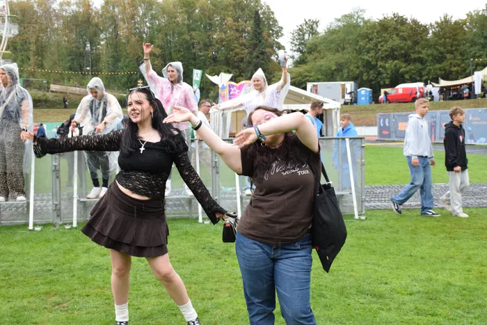 Zwei Frauen tanzen ausgelassen auf einer Wiese, während im Hintergrund weitere Besucher in Plastikregencapes feiern.