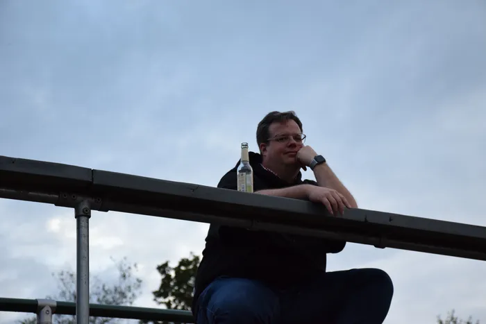 Ein Mann mit Brille lehnt an einem Metallgeländer, hält eine Flasche und stützt sein Kinn auf seine Hand.