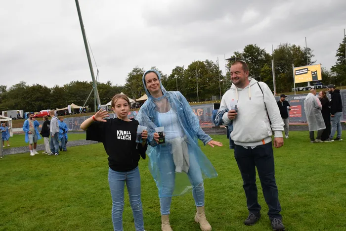 Ein lächelndes Mädchen, eine Frau in einem blauen Regenponcho und ein Mann im weißen Hoodie stehen auf einer Rasenfläche.