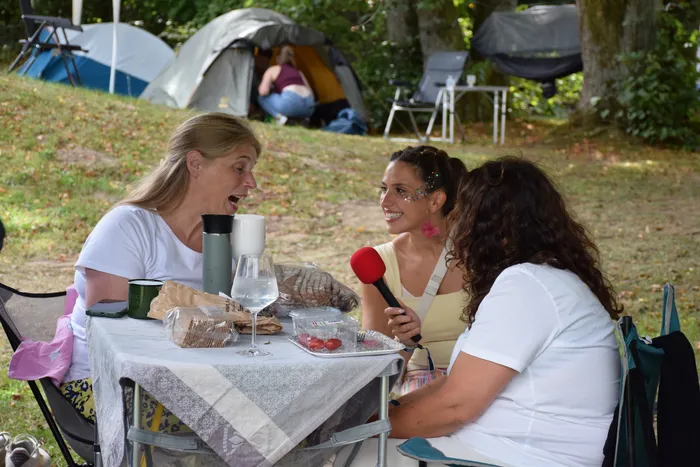 Drei Frauen unterhalten sich an einem Tisch im Freien, eine hält ein rotes Mikrofon. Im Hintergrund stehen Zelte auf einer Wiese.
