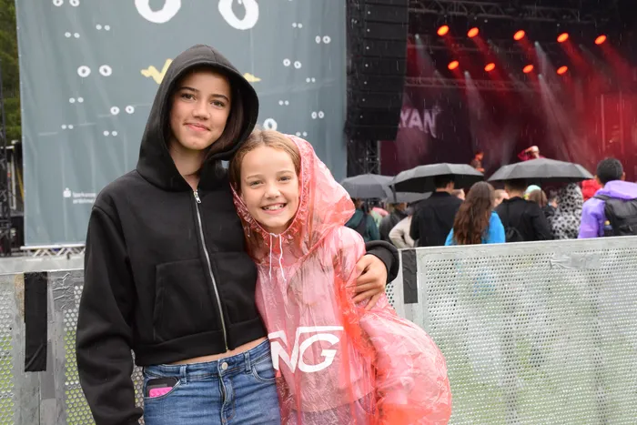 Zwei lächelnde Mädchen, eines in einem schwarzen Kapuzenpullover und eines in einem rosa Regenmantel, posieren vor einer beleuchteten Bühne im Regen.