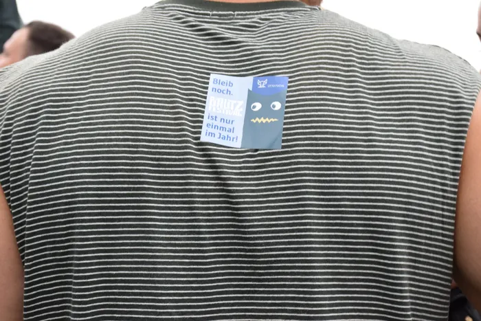 Blauer Aufkleber auf einem grau-weiß gestreiften Shirt mit der Aufschrift „Bleib noch. BAUTZ FESTIVAL ist nur einmal im Jahr!“.