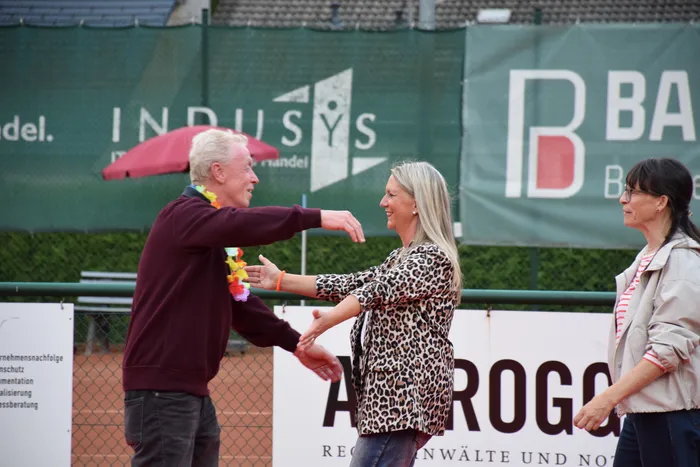 Ein Mann mit einer Blumenkette und eine Frau in einem Leopardenmuster-Oberteil begrüßen sich auf einer Tennisanlage.