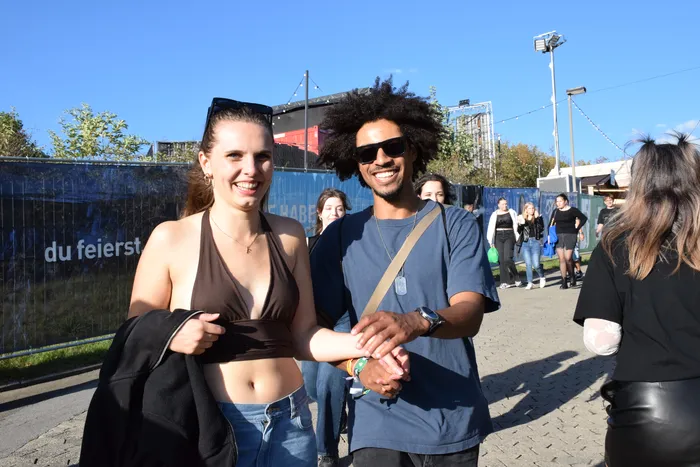 Eine lächelnde junge Frau in einem braunen Top und ein junger Mann mit Afro und Sonnenbrille halten Händchen bei sonnigem Wetter.