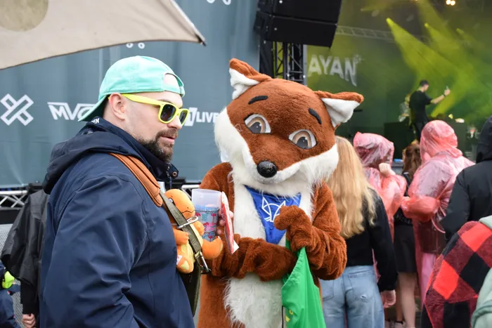 Ein Mann mit hellgrüner Kappe und gelber Sonnenbrille steht neben einem braun-weißen Fuchs-Maskottchen bei einer Veranstaltung im Freien.