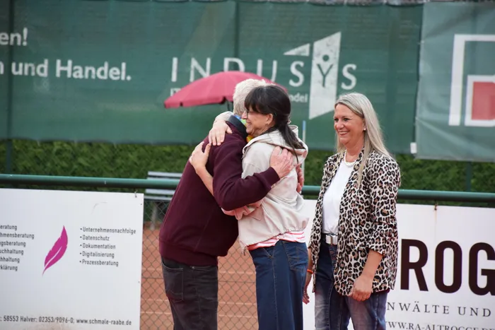Zwei Personen umarmen sich auf einem Tennisplatz, während eine Frau im Leopardenmuster-Sakko daneben steht und lächelt.