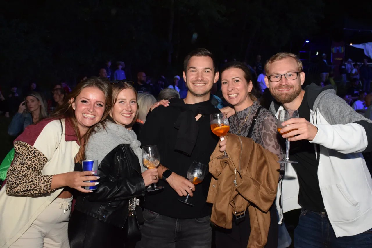 Fünf lächelnde Personen halten Gläser mit Getränken in die Kamera bei einer Abendveranstaltung.