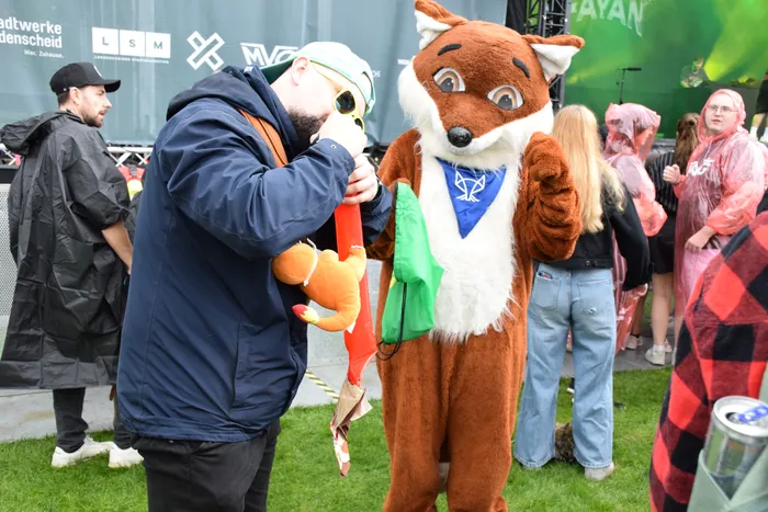 Ein Mann in blauer Jacke steht neben einem Fuchs-Maskottchen auf dem Bautz-Samstag; im Hintergrund tragen Besucher Regenponchos.